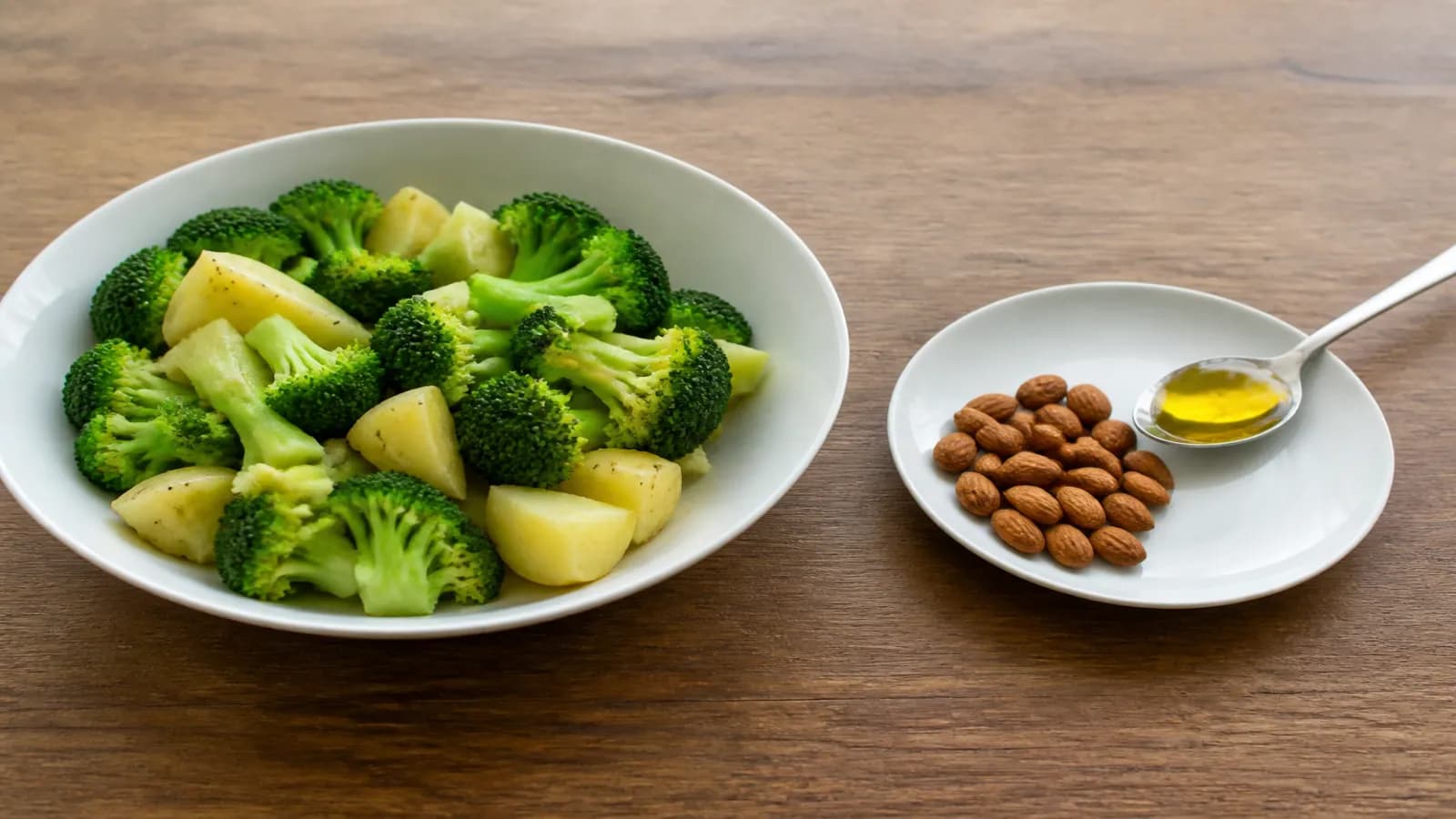 Grande assiette de brocoli et pomme de terre à côté d’une petite poignée d’amandes et d’une cuillère d’huile, table en bois.